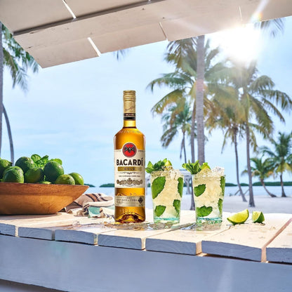 Garrafa de rum Bacardi Carta Oro ao lado de dois drinks com limão e hortelã em cenário tropical.
